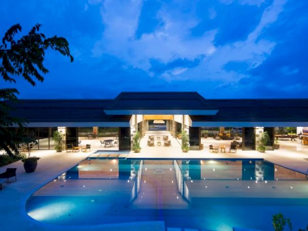 A luxurious, modern building with an illuminated swimming pool in front, surrounded by lounge chairs and tables under a twilight sky.