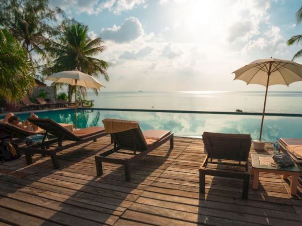 A serene poolside scene with deck chairs, umbrellas, and coconut trees overlooking a calm ocean under a partly cloudy sky, bathed in sunlight.