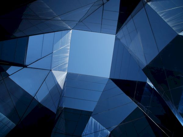 The image shows several reflective and modern glass buildings converging towards the center, forming a geometric pattern with a clear sky visible in the middle.