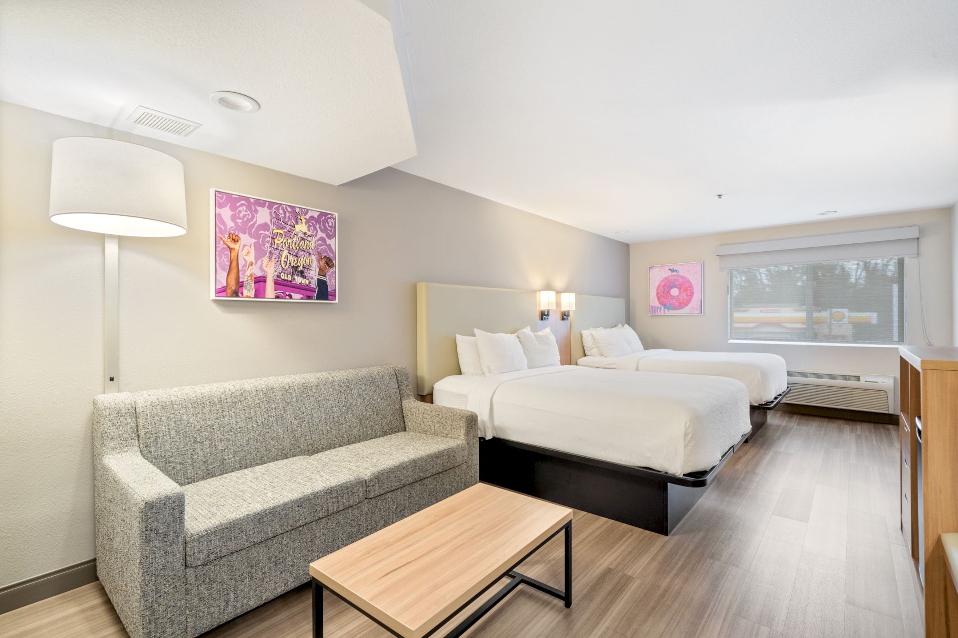 The image shows a modern hotel room with two beds, a gray sofa, a wooden coffee table, a wall lamp, and colorful artwork on the walls.