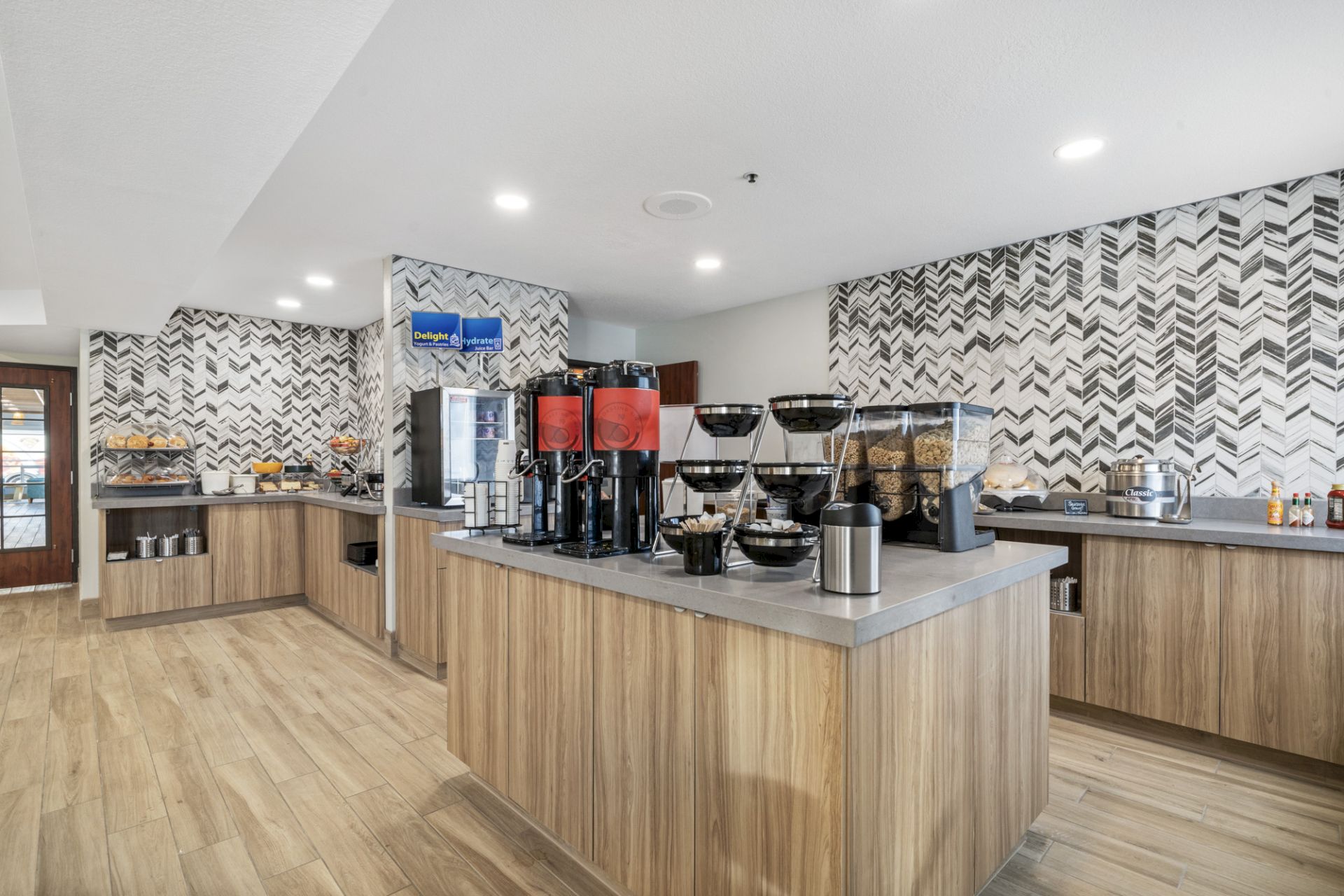 This image shows a modern self-service breakfast area with coffee dispensers, cereal dispensers, and various food items on a wood-themed countertop.