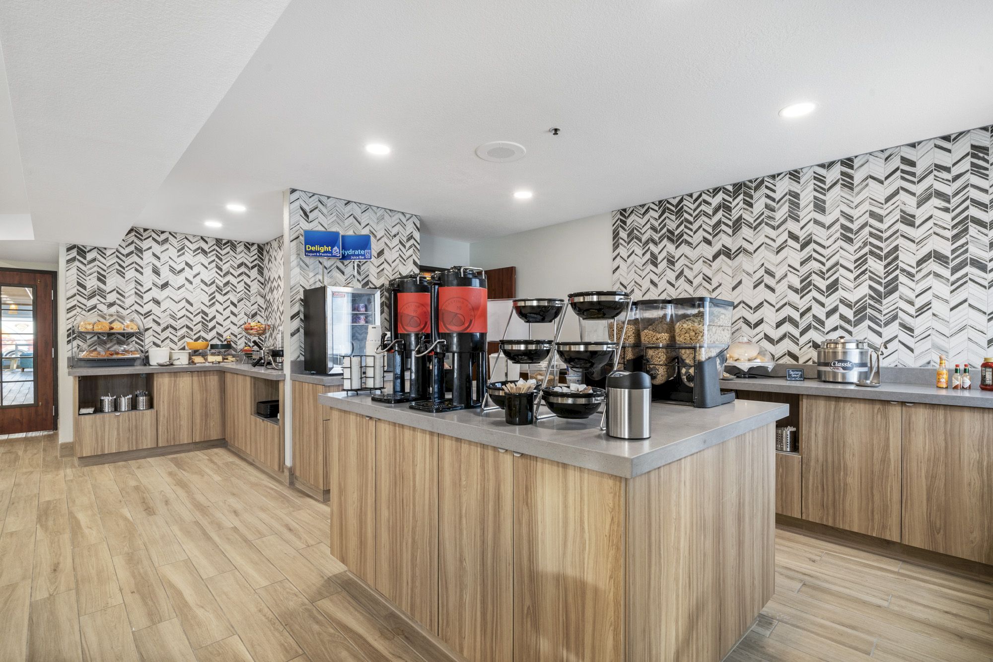 This image shows a modern self-service breakfast area with coffee dispensers, cereal dispensers, and various food items on a wood-themed countertop.