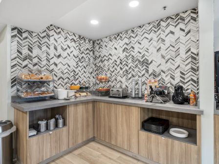 A modern kitchen corner setup with various appliances, bread, fruit, toaster, and a stylish herringbone backsplash, neatly organized and well-lit.