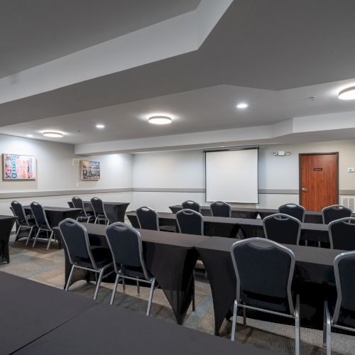 The image shows a conference room with rectangular tables, black chairs, a projector screen, and some artwork on the walls.