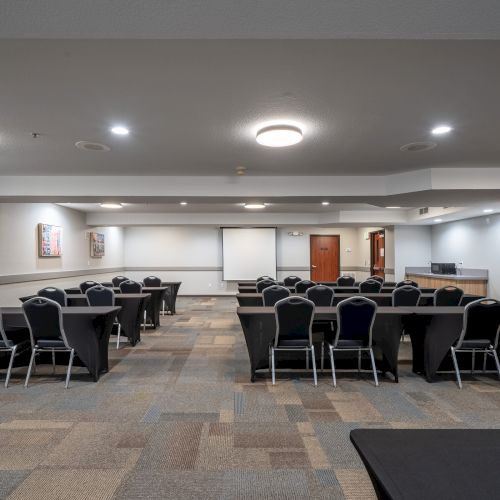 A conference room with multiple round tables, chairs, a projector screen, and some artwork on the walls. There are stacked chairs in the corner.