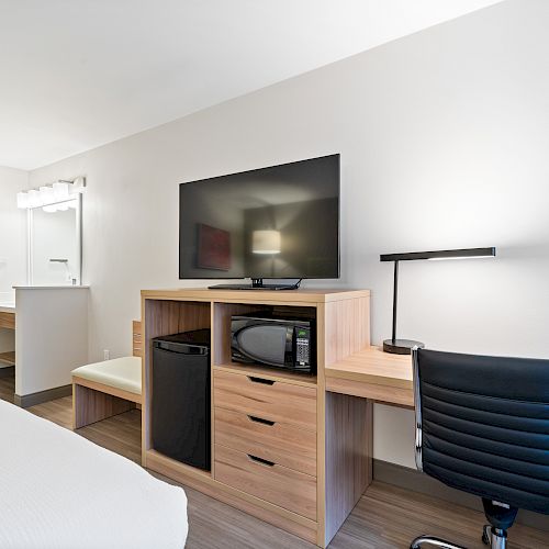 The image shows a hotel room with a bed, desk, office chair, TV, microwave, mini-fridge, and a lit vanity area near the entrance.