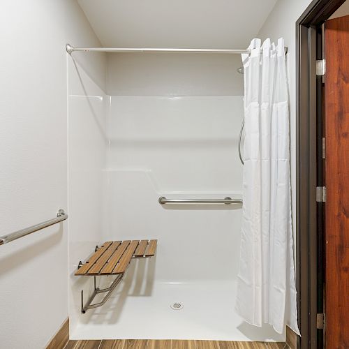 The image shows an accessible shower with a foldable bench, grab bars, white curtain, and a wooden door partly open.