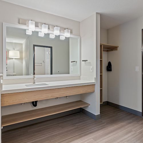 A modern bathroom with a large mirror, illuminated by lights above the sink, a wooden shelf beneath, and a closet area to the right ending the sentence.