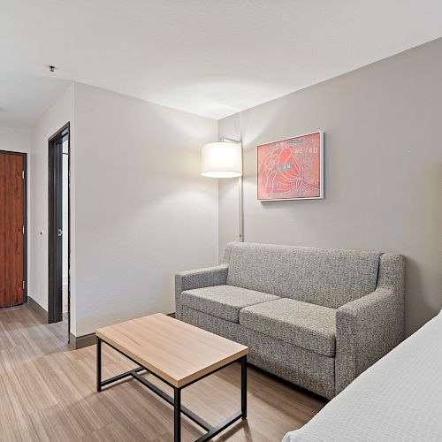 A modern hotel room features a sofa, coffee table, bed, wall art, and a door with wooden flooring and neutral decor.