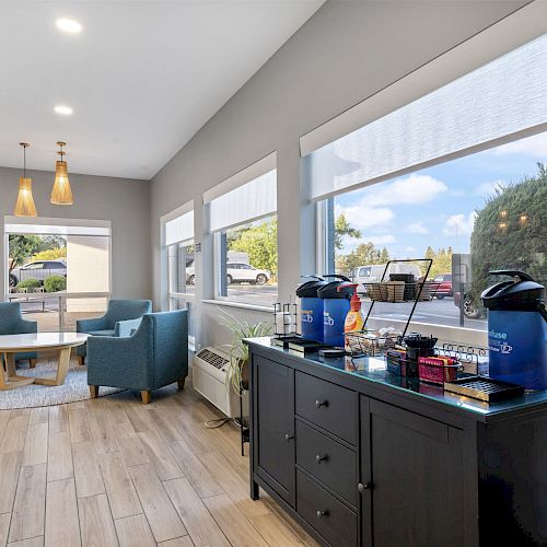 A cozy room with seating, a coffee station on the right, large windows, wooden floor, and hanging lights, creating a welcoming atmosphere.