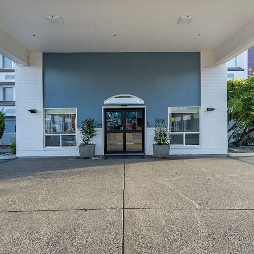 The image shows the entrance to a building with double glass doors, flanked by two potted plants and framed by a canopy.