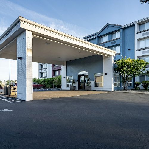 The image shows the entrance of a modern hotel with a covered driveway, glass doors, and surrounding greenery under clear skies.