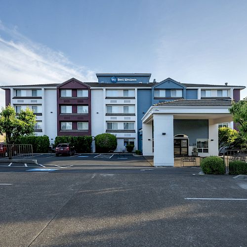 A four-story hotel with red, white, and blue exterior sections, trees, a parking lot with minimal cars, and an entrance cover.