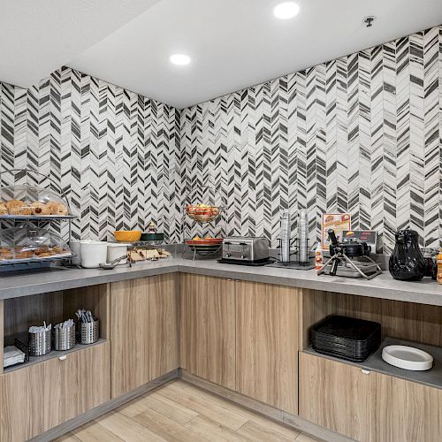 This image shows a modern kitchen or breakfast nook with a variety of equipment, including a toaster, coffeemaker, and assorted food items on the counter.