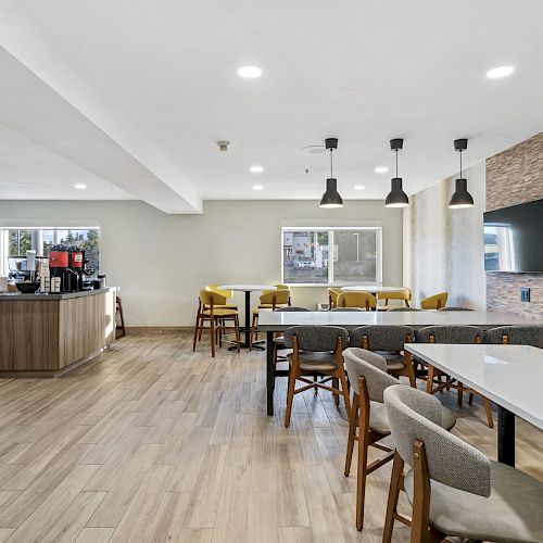 A bright, modern dining area features several tables and chairs, a coffee station, pendant lighting, wooden flooring, and a wall-mounted TV.