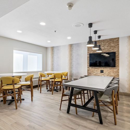 A modern dining area with several tables and chairs, pendant lighting, a wall-mounted TV, and a mixture of seating styles, including a booth.