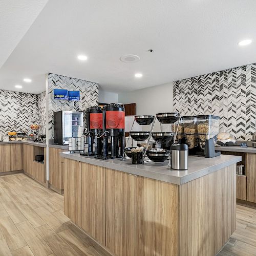 This image shows a breakfast buffet area with coffee dispensers, cereal dispensers, a waffle maker, and various kitchen appliances on wooden counters.