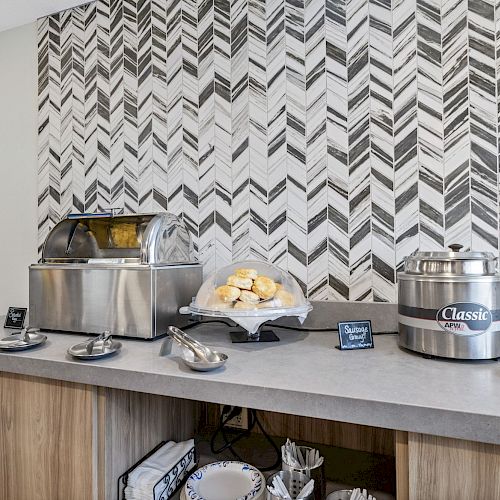 The image shows a breakfast buffet setup with a waffle maker, pastries, soup container, cups, and napkins on a countertop against a patterned wall.