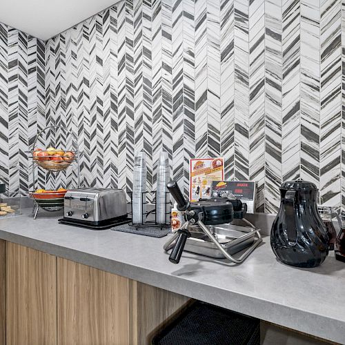 This image shows a kitchen counter setup with appliances like a toaster, waffle maker, coffee maker, and items such as a bowl of fruit and syrup.