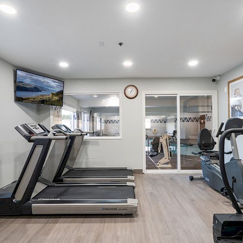 A modern gym with treadmills, an exercise bike, a wall-mounted TV, a clock, and large mirrors reflecting the equipment and surroundings.