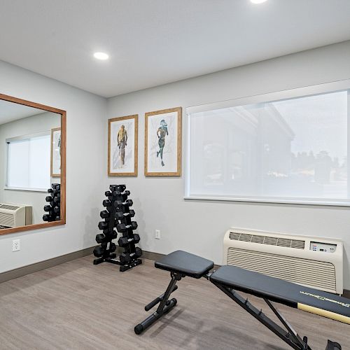 This image shows a small home gym with a bench, a dumbbell rack, and a large mirror. The room has framed pictures and a window with a roller shade.