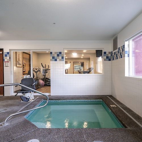 This image shows an indoor pool area with a small pool, exercise equipment visible through a window, and a door leading to another room.