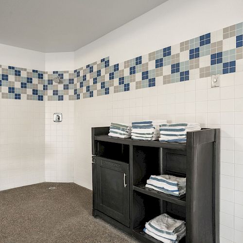 The image shows a tiled wall with blue accents, a wooden cabinet with folded towels, a chair, and a wooden door with a silver handle.