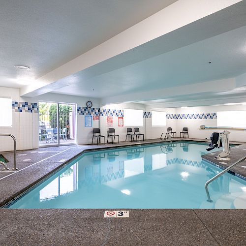 This image shows an indoor swimming pool area with chairs along the walls, a hot tub, and a pool lift near the entrance.