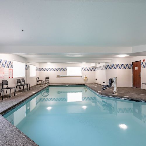 An indoor swimming pool with several chairs along the wall, a clock, and safety signs posted. The room has tiled walls and a door on the right.