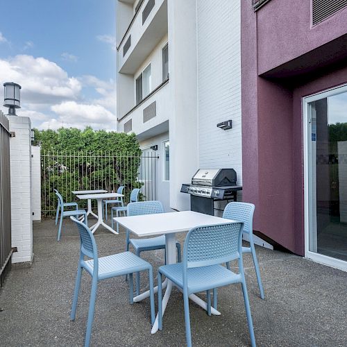 An outdoor patio with light blue chairs and tables, a barbecue grill, and a fence surrounding the area next to a building.