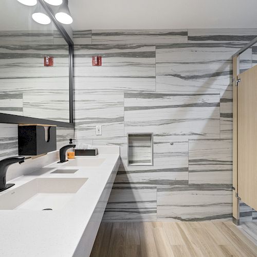 A modern public restroom with a sleek countertop with sinks, soap dispenser, large mirror, and a stall with a wooden door and tiled walls.