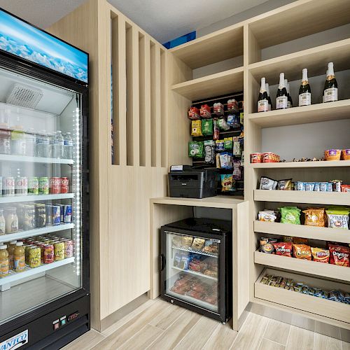 A small convenience area with a fridge stocked with drinks, shelves of snacks, various bottled beverages, a small beverage cooler, and a microwave.