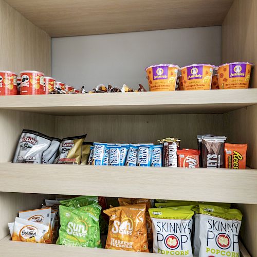 The image shows a cupboard with various snacks, including instant noodles, protein bars, and bags of popcorn and chips on three shelves.