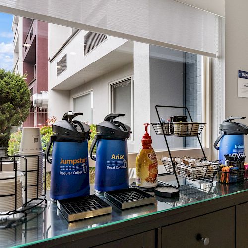 A coffee station with coffee dispensers, cups, lids, creamers, and condiments is set up against a window with a view of a building and greenery outside.