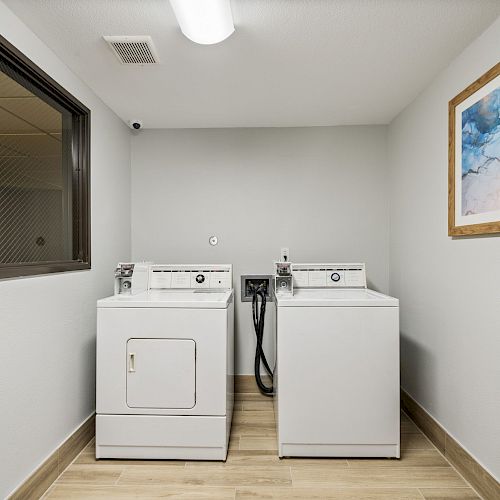 The image shows a laundry room with a white washer and dryer set, a small window, and a framed abstract painting on the wall.