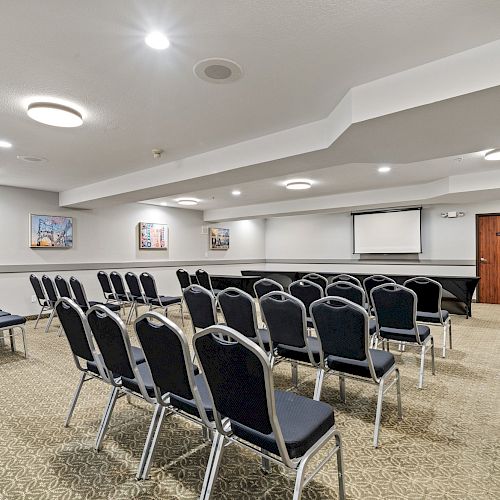 The image shows a conference room with rows of black chairs, a projector screen, framed artwork on the walls, and an American flag near the door.