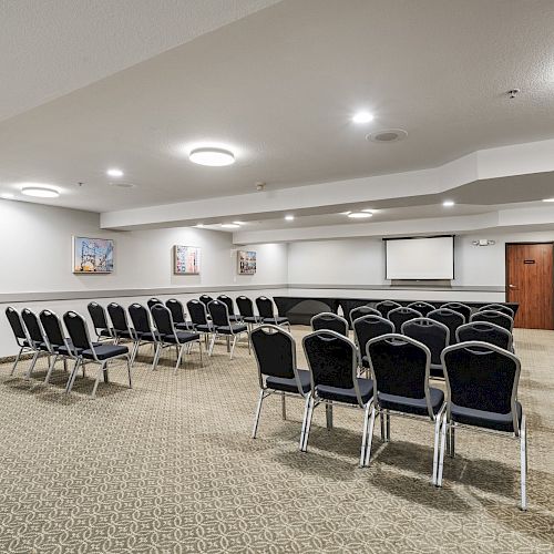 A conference room features rows of chairs, a projector screen, artwork on the walls, and a carpeted floor under bright lighting.