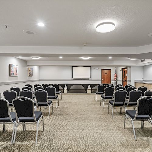 A conference room with rows of chairs facing a projector screen, decorated with wall art, and extra chairs stacked in the corner.