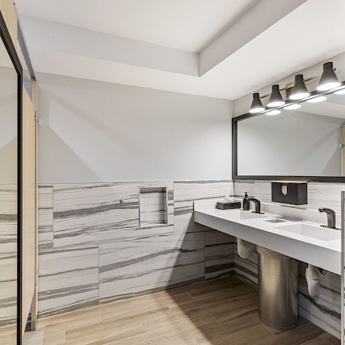 The image shows a modern bathroom with a large mirror, dual sinks, ample lighting, marble walls, and a wooden floor, maintaining a minimalist style.