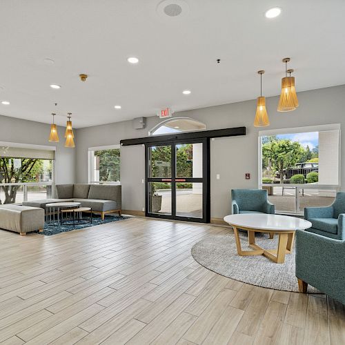 A modern, spacious lobby with large windows and natural light. The area features grey sofas, blue chairs, and wooden tables.