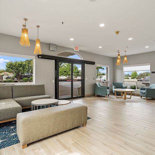 A modern lobby with seating areas, pendant lights, large windows, and a glass entrance door, featuring neutral and blue decor.