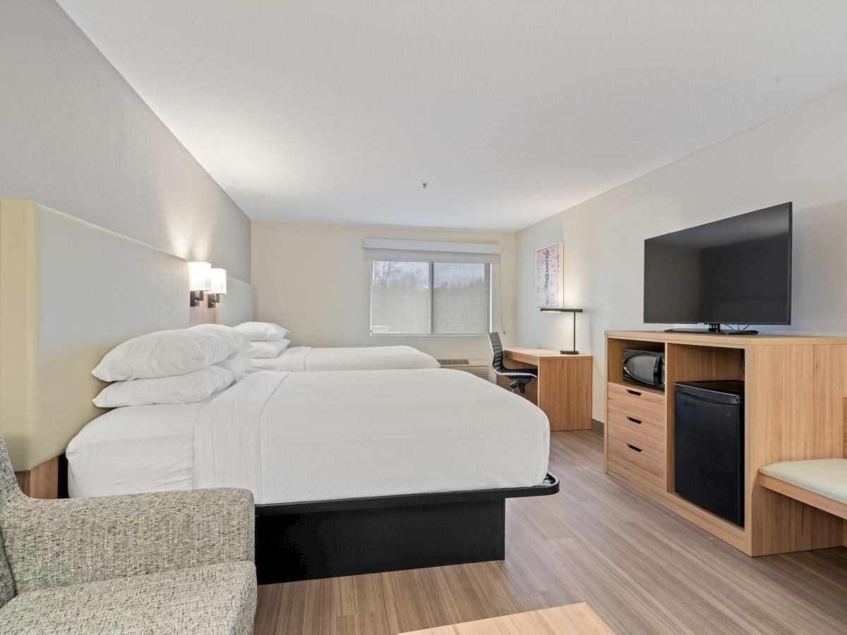 This image depicts a hotel room with two beds, a TV on a wooden stand, a desk, a chair, and a window with a view outside.