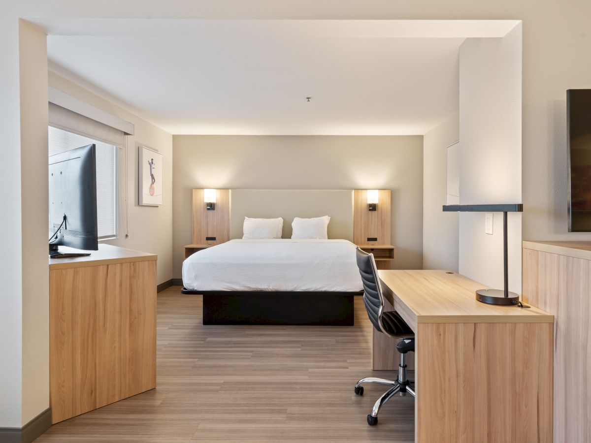 A modern hotel room features a bed, nightstands, desks, chairs, and a TV. The decor is minimalist with light wood accents, and the room is well-lit.