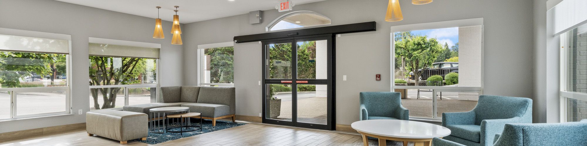 A modern, spacious lobby with large windows, seating area, hanging lights, blue chairs, and wooden flooring, leading to glass doors.