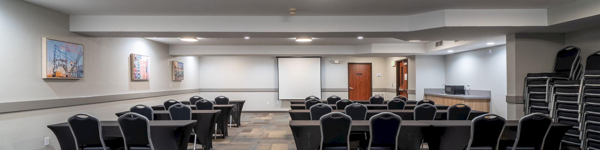 An empty conference room with rows of black chairs and tables, a projector screen at the front, and stacked chairs on the right side.