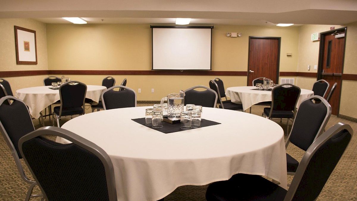 A conference room with round tables covered in white cloth, chairs, a projector screen, and a table setup with glasses and water pitchers.