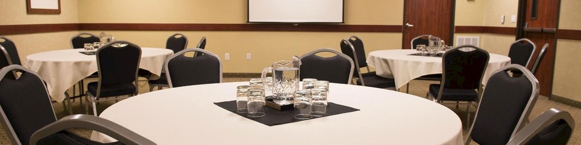 A conference room with round tables covered in white cloth, chairs, a projector screen, and a table setup with glasses and water pitchers.