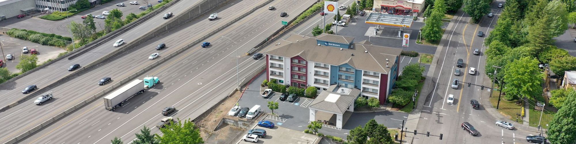 An aerial view of a highway with multiple lanes and vehicles, nearby buildings, a gas station, and surrounding greenery.