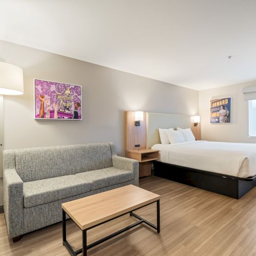 The image shows a modern hotel room with a bed, grey couch, small table, wall art, floor lamp, and window with a blind, all in neutral tones.