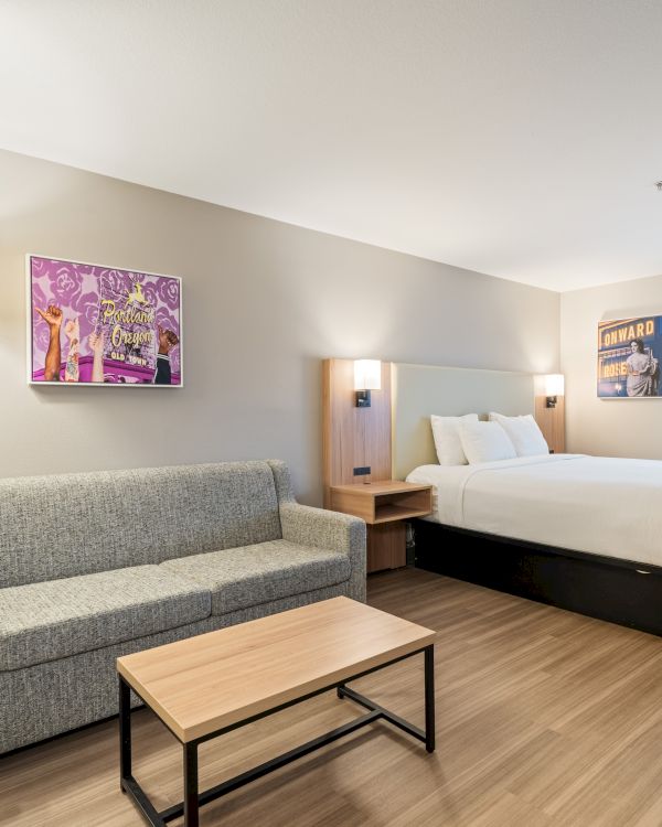 The image shows a modern hotel room with a bed, grey couch, small table, wall art, floor lamp, and window with a blind, all in neutral tones.